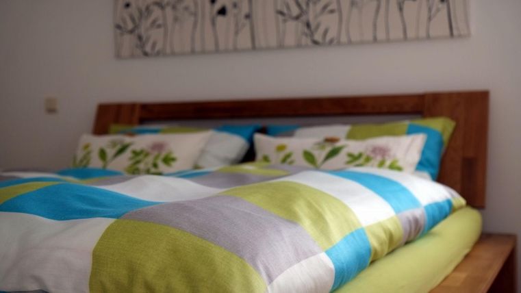 Ein gemütliches Bett mit bunten Bettwäsche in Blau und Grün. An der Wand hängt ein schlichtes Kunstwerk mit floralen Motiven.