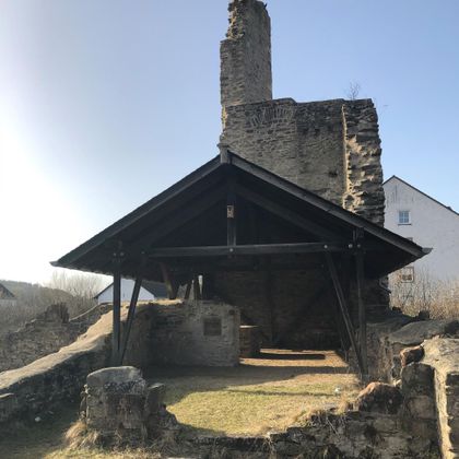 Im Hintergrund sieht man eine hochaufragende etwa 5 m breite Mauer. An deren rechter Seite setzt eine im rechten Winkel noch höhere Wand aus Bruchsteinen an.
Vor der Wand hat man eine Holzkonkruktion als Dach errichtet, die an der hinteren Wand abschließt. Sie ist an beiden Seiten und nach vorne offen. Ganz im vordergrund sieht mann einige Bauerreste, die teilweise mit Gras bewachsen sind. In den  Flächen zwischen den Mauernresten hat sich ebenfalls Gras ausgebreitet.