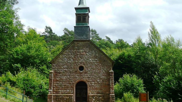 Die Büschkapelle in Gerolstein, eine kleine Steinkapelle mit Turm, umgeben von Wald und Bänken, unter einem bewölkten Himmel.