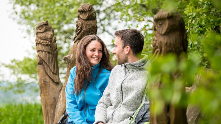 Ein Paar sitzt auf einer Bank mit Eulenschnitzereien im Freien.