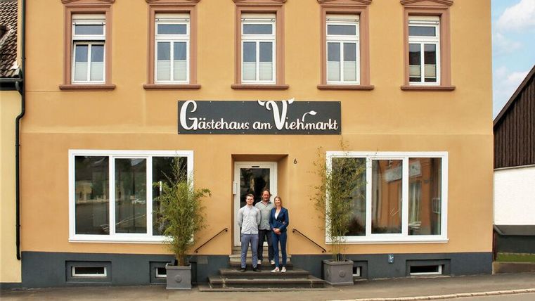 Une maison d'hôtes avec deux personnes devant l'entrée. La façade est de couleurs chaudes et possède de grandes fenêtres.