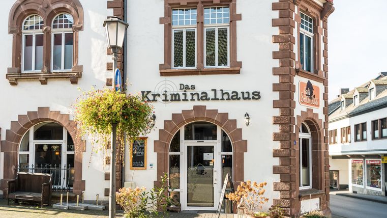 Eingang des Kriminalhauses in Hillesheim, dekoriert mit Pflanzen. Rechts ist das Café Sherlock zu sehen. Historische Architektur mit roten Akzenten.