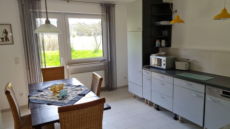 A bright kitchen with a wooden dining table and two chairs. In the background, there is a large window front overlooking a green garden.