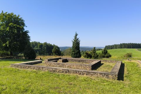 Ruinen eines alten Gebäudes auf einer grünen Wiese, umgeben von Bäumen und Hügeln unter klarem, blauem Himmel.