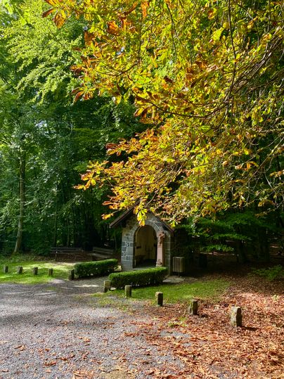 Eine kleine Kapelle am Wegesrand umgeben von Laubbäumen.