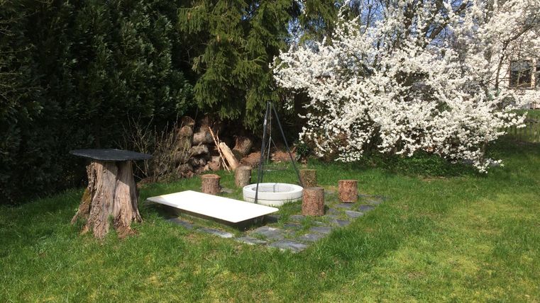 Eine grüne Wiese mit einem weißen Tisch, einem runden Feuerplatz und Sitzstämmen aus Holz. Im Hintergrund blüht ein weißer Baum.