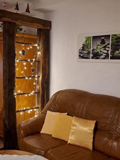 Eine gemütliche Wohnzimmerecke mit einem braunen Sofa und Kissen. Im Hintergrund hängen Lichter und eine Dekoration mit Pflanzenbildern ist an der Wand zu sehen.