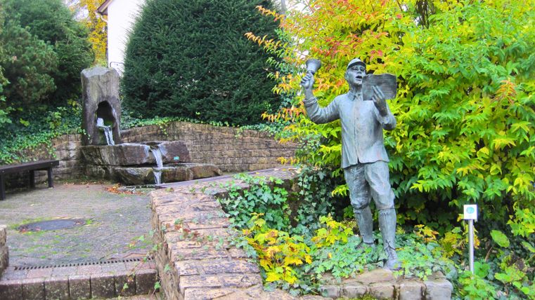 Statue eines Mannes mit Glocke und Papier in einem Garten mit Brunnen und Pflanzen.