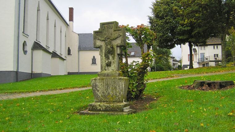 Steinkreuz vor einer Kirche mit Bäumen im Hintergrund.