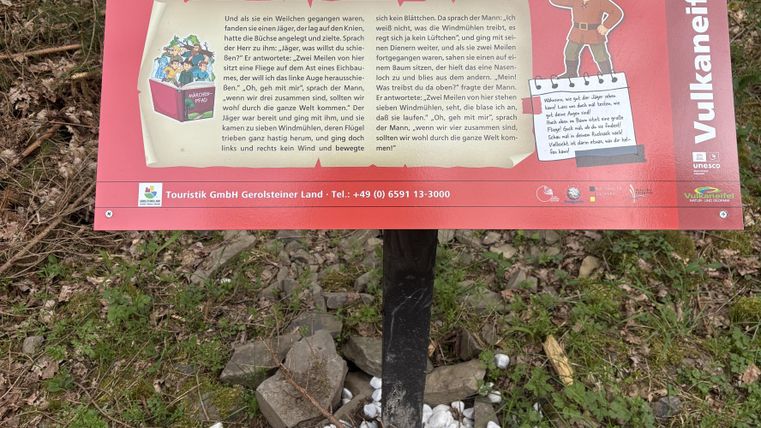 Ein Informationsschild über den Märchenpfad in Jünkerath. Es enthält eine Geschichte und ist von einer grünen Umgebung umgeben.