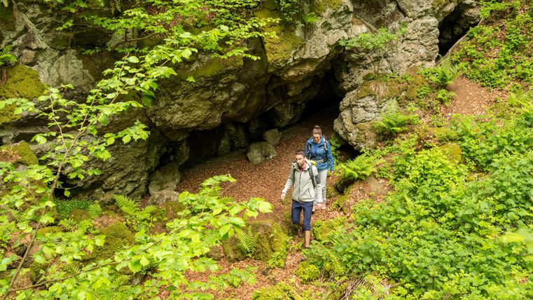 Deux randonneurs passent devant une grotte rocheuse sur un sentier forestier verdoyant. Une végétation luxuriante entoure le chemin.