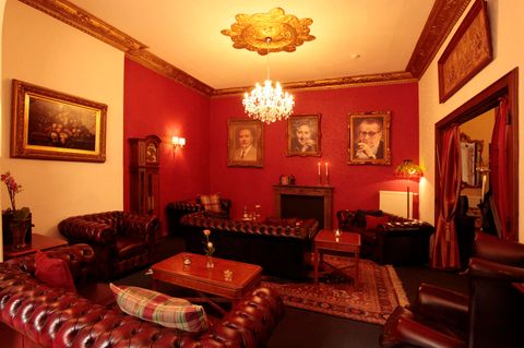 Un salon élégant avec des murs peints en rouge et des meubles en cuir confortables. Des portraits historiques sont accrochés aux murs et un plafonnier élégant crée une ambiance lumineuse.