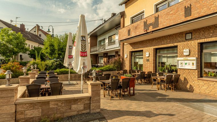 Terrasse des Restaurants mit Tischen, Stühlen und Sonnenschirmen. Die Umgebung ist grün und das Wetter sonnig.