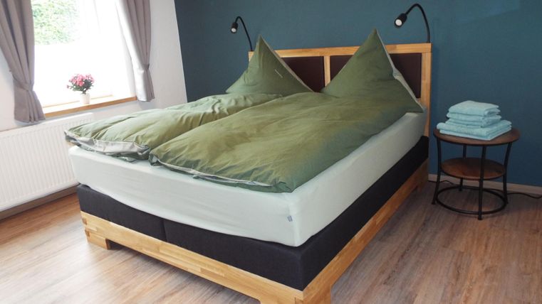 Ein gemütliches Schlafzimmer mit einem großen Bett und grünen Bettdecken. Im Raum gibt es einen Nachtisch und helle Vorhänge.