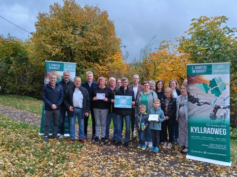18 Personen stehen vor herbstlichen laubbäumen auf einer schmalen asphaltierten Straße zusammen zwischen zwei Bannern für den Radaktionstag "Grenzenlos Kyllradweg" und lächeln in die Kamera. Drei Personen halten stolz jeweils einen Gewinn in den Händen.