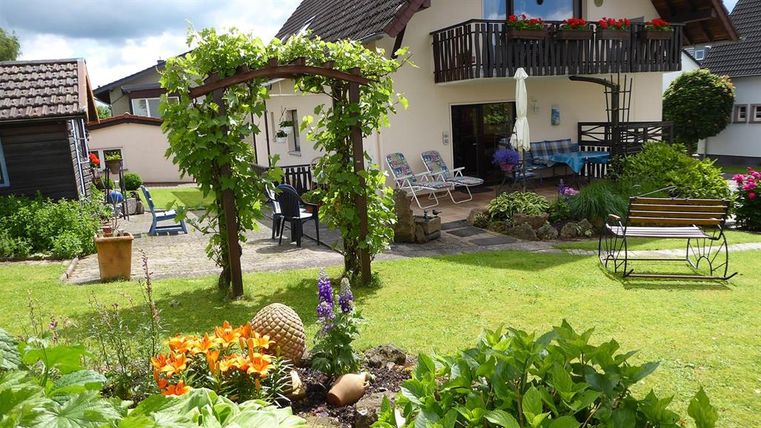 Ein schöner Garten mit bunten Blumen und einer Laube aus Weinreben. Im Hintergrund steht ein Haus mit Balkon und Gartenmöbeln.