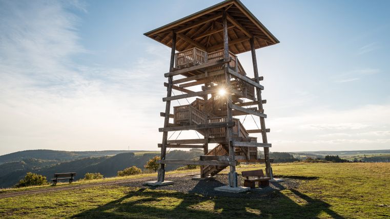 Een houten uitkijktoren op een weiland met uitzicht op een heuvelachtig landschap bij zonsondergang.