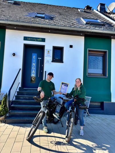 Zwei Personen stehen mit Fahrrädern vor einem Gebäude mit der Aufschrift „harmony café“. Es ist ein sonniger Tag und die Atmosphäre wirkt einladend.