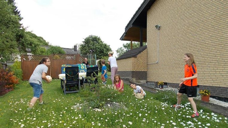A group of children is playing in the garden, surrounded by flowers and grass. In the background, a house can be seen.