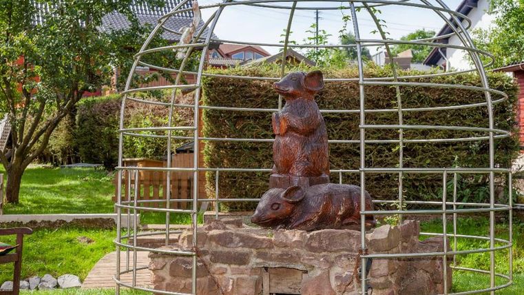Eine Skulptur von zwei Mäusen in einem Metallgitter. Die Figuren sind auf einem Steinfundament in einem grünen Garten platziert.