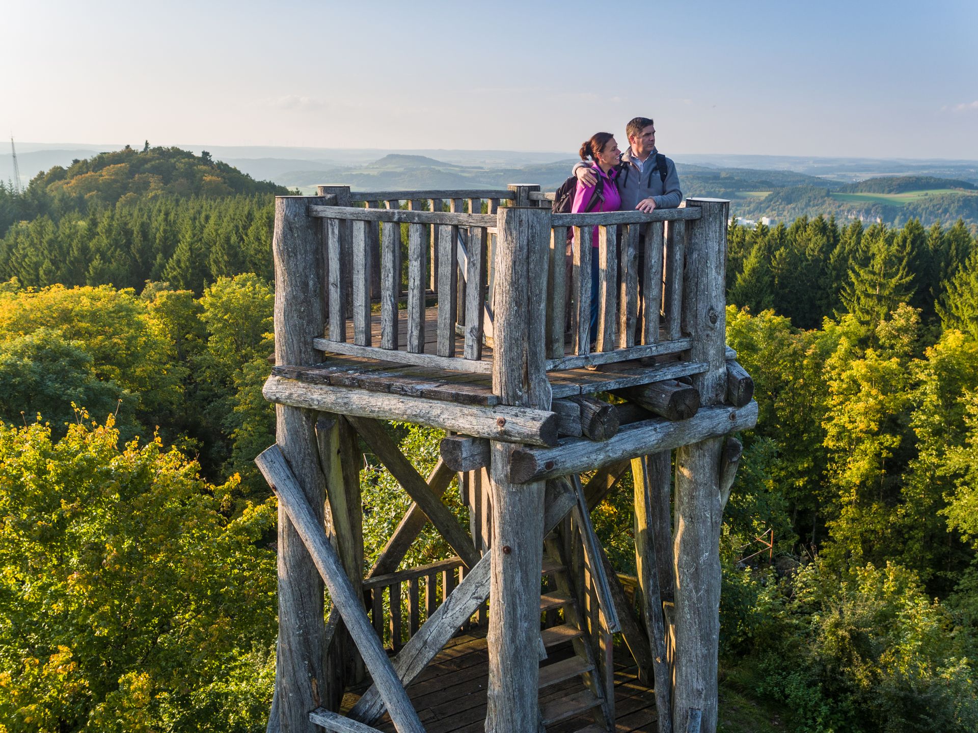 zwei Personen auf einem Holzaussichtsturm inmitten eines herbstlichen Waldes mit weitem Blick über die Landschaft.
