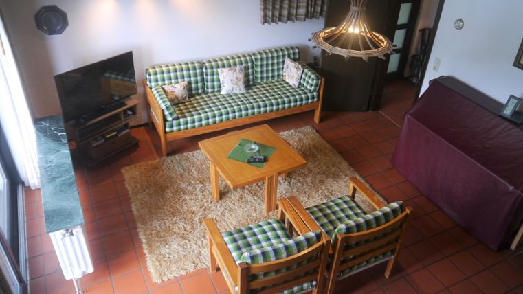 Ein gemütlicher Wohnraum mit einem grünen Couchset und einem Holzkaffee-Tisch. Der Raum verfügt über einen Fernseher und ein warmes Licht von einer Deckenlampe.