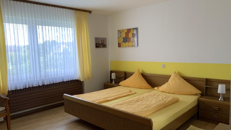 Ein helles Schlafzimmer mit einem großen Doppelbett und gelben Kissen. Die Fenster sind mit Vorhängen versehen und es gibt einen Holzfußboden.