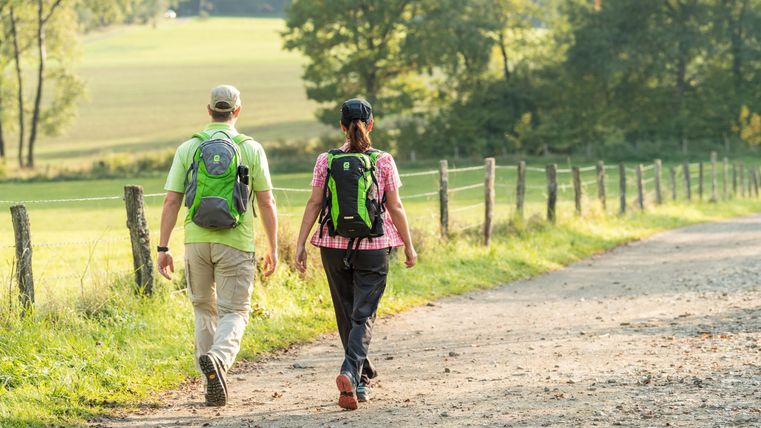 Un couple marche sur un chemin de campagne, entouré d'arbres et de prairies. Tous les deux portent des sacs à dos et profitent de la nature.