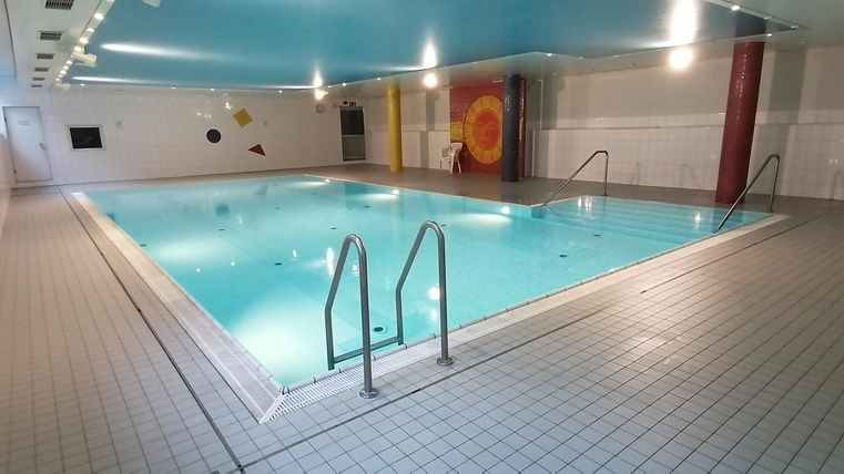 Ein Lehrschwimmbecken mit blauer Decke, bunten Wanddekorationen und zwei Einstiegsleitern. Die Wände sind weiß gefliest.