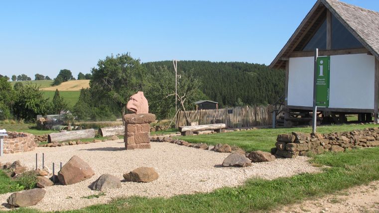 Freilichtmuseum mit Skulptur auf Steinsockel, Holzhütte und Infotafel. Umgeben von Wiesen und Bäumen unter klarem Himmel.