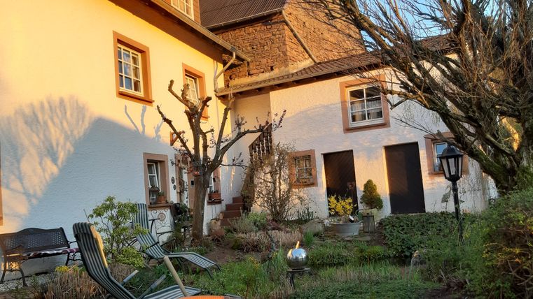 Een charmant huis met witte muren en grote ramen. De tuin is groen, met bomen en een gezellige zitgelegenheid.