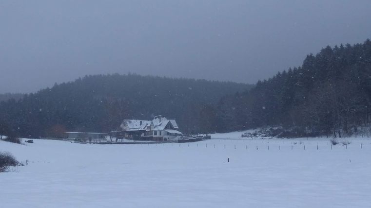 Un paysage enneigé avec une grande maison en arrière-plan. Le ciel est gris et nuageux.