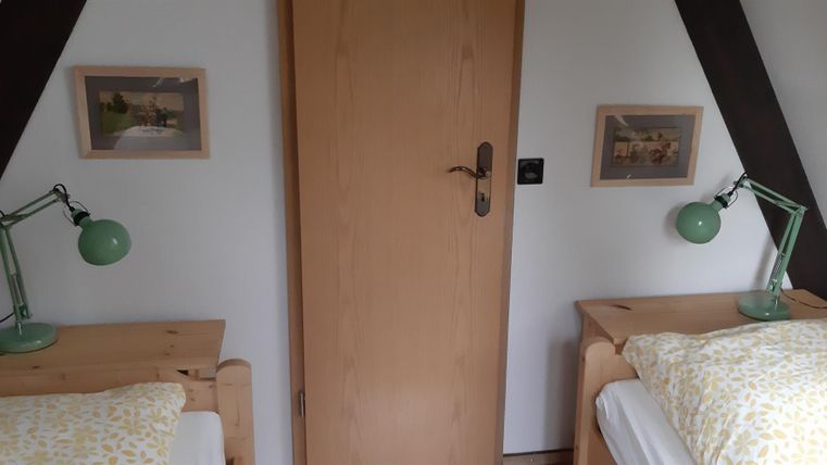 Ein gemütliches Schlafzimmer mit zwei Einzelbetten aus Holz. An den Wänden hängen Bilder, und auf jedem Nachttisch steht eine grüne Lampe.