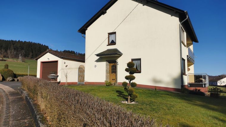 A modern house with a bright paint job and a well-kept garden. In the background, gentle hills and a clear sky can be seen.