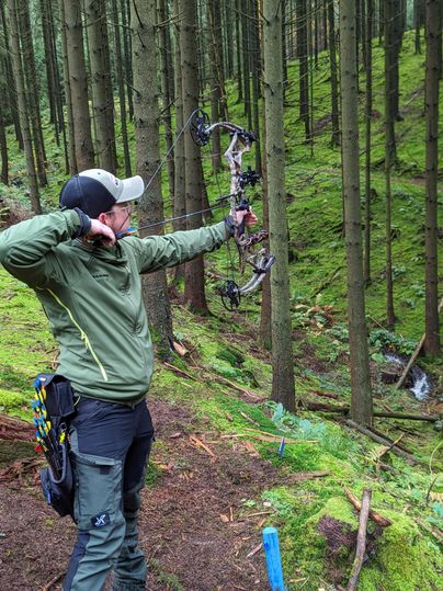 Un homme se tient dans la forêt et vise avec un arc. Autour de lui se trouvent de grands arbres et de la mousse verte.