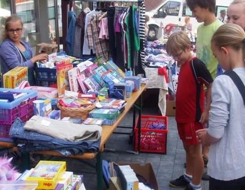 Bunter Flohmarktstand mit Kleidung, Büchern und Spielzeug. Einige Personen stehen davor und betrachten die Ware.
