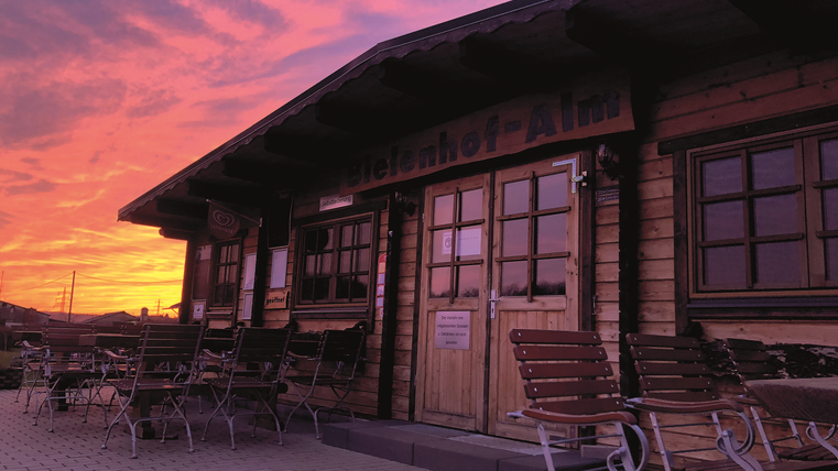 Terrasse der Bielenhof Alm mit Holzbänken vor dem Holzgebäude. Bunter Himmel durch die untergehende Sonne im Hintergrund.