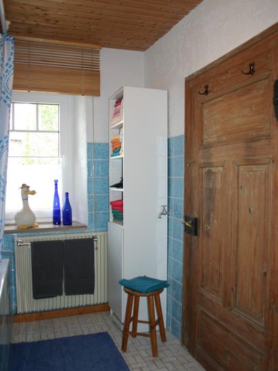 Een lichte badkamer met blauwe tegels en houten details. Een kast met kleurrijke handdoeken en een houten stoel zijn zichtbaar.