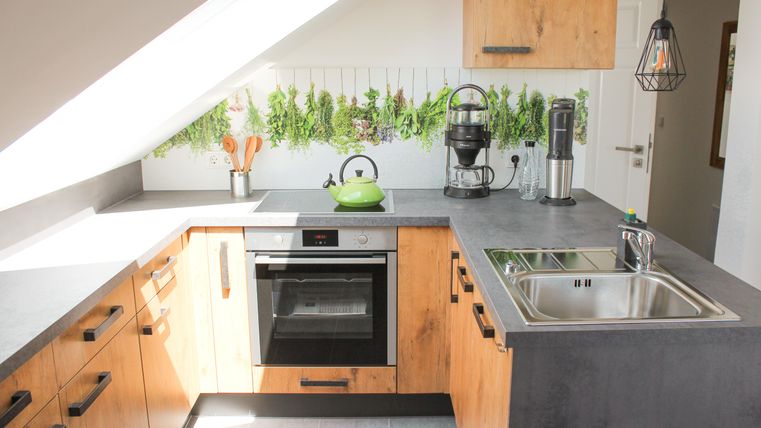 Eine moderne Küche mit Holzschränken und einer klaren Arbeitsfläche. Über der Spüle hängen frische Kräuter in Pflanzenbehältern.