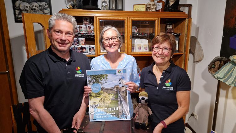 Drei Personen präsentieren ein Plakat für den Hillesheimer Detektiv-Trail in einem Raum mit dekorativen Gegenständen.