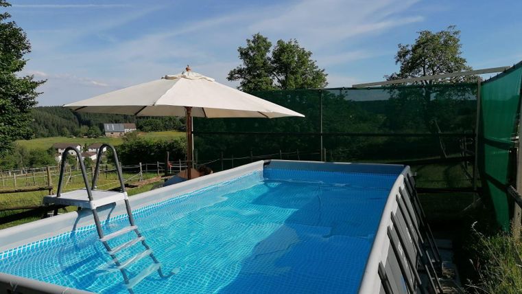 Een opzetzwembad met helder water en een trap. Naast het zwembad staat een parasol en op de achtergrond zijn bomen en gras te zien.