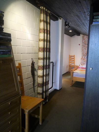 Ein einfacher Flur mit weißen Wänden und Holzdecke. Auf der linken Seite stehen einige Stühle und es gibt einen Blick auf ein Schlafzimmer am Ende des Flurs.