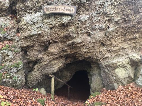 Eingang zur Martins-Höhle in Hohenfels-Essingen, umgeben von Laub und Felsen. Ein Holzschild mit der Aufschrift 'Martins-höhle' ist sichtbar.