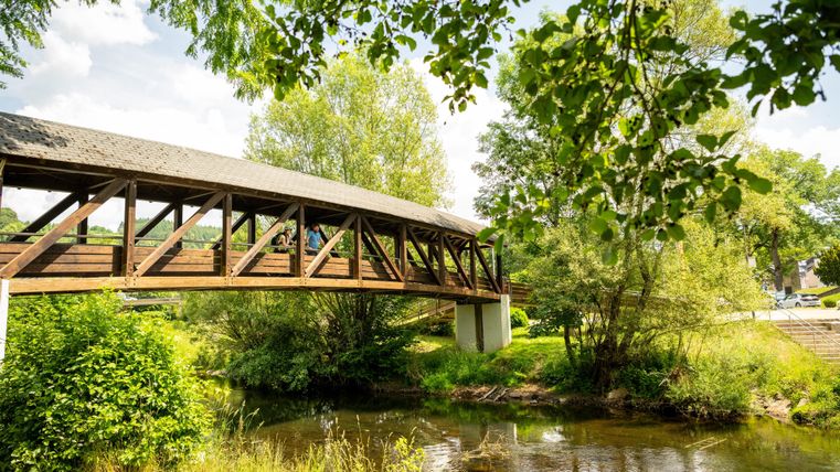 Eine malerische Holzbrücke über einen ruhigen Fluss. Üppiges Grün umgibt die Szene, und der Himmel ist teilweise bewölkt.