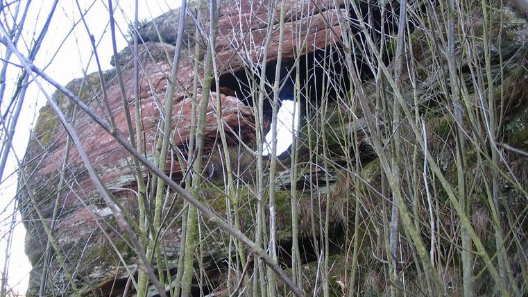 Eine erodierte Felsformation, umgeben von dünnen Ästen und Pflanzen. Im Hintergrund ist ein Fenster in der Felswand sichtbar.