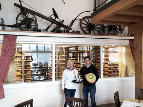 Ein Mann und eine Frau stehen vor eine großen Glasscheibe, hinter der in Regalen zahlreiche Käselaibe reifen. Dei Frau trägt ein Tablett mit Getränken und der Mann einen Käselaib.