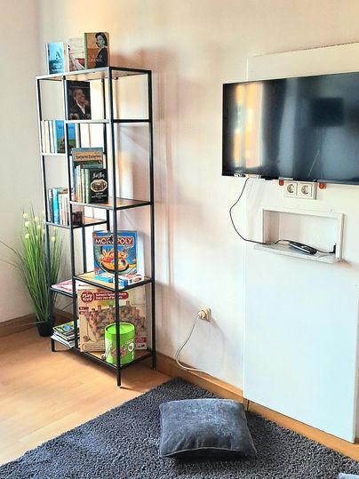 Ein gemütlicher Raum mit einem Bücherregal und einem Fernseher. Auf dem Boden liegt ein weicher Teppich, und eine Pflanze verleiht dem Raum Frische.