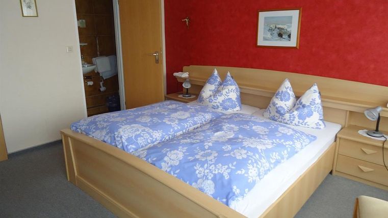 A cozy bedroom with a double bed and blue bedcovers. The walls are painted red, and there is a nightstand with a lamp.