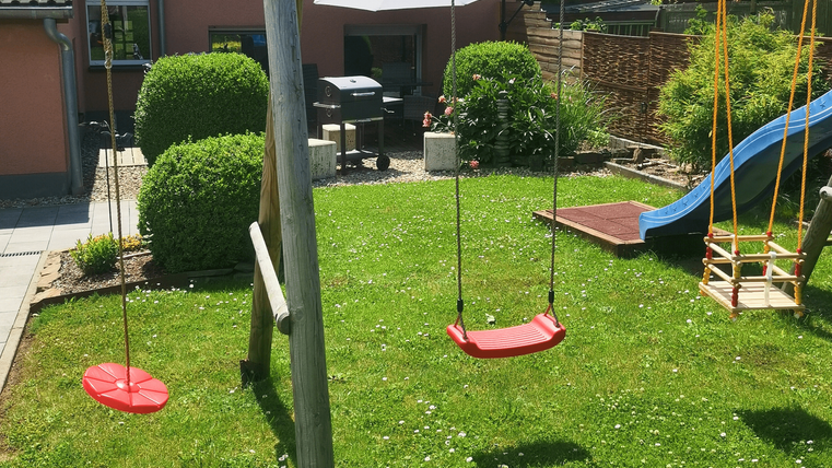 Ein gepflegter Garten mit zwei Schaukeln, einem Rutschen und einem Sonnenschirm. Im Hintergrund sind Grüne Büsche und ein Grill zu sehen.