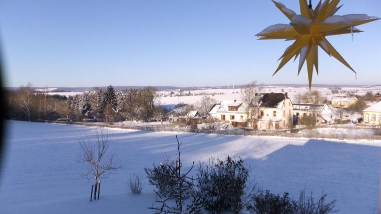 Eine winterliche Landschaft mit einer schneebedeckten Wiese und Häusern im Hintergrund. Im Vordergrund hängt ein dekorativer Stern.